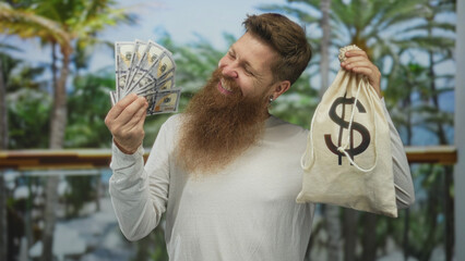 Man holds money bag and fan of cash with bare hands while smiling among palm trees in forest;...