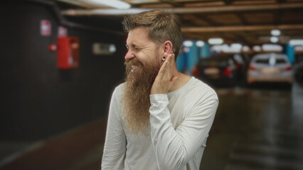 Man with bushy beard and casual white shirt holds hand to ear in building filled with blurred cars...
