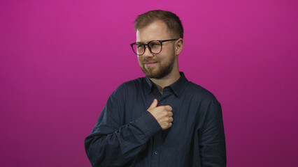Man with glasses holding hand to chest in studio on bright magenta backdrop, wearing navy shirt and...