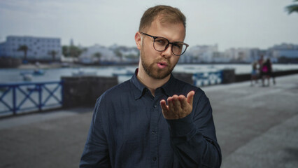 Man blows a kiss with open hand on seaside street promenade near harbor while wearing glasses and beard; playful flirtation.