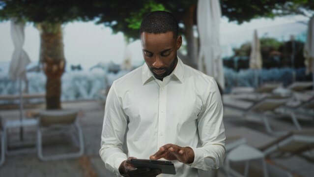 Man in a white shirt tapping a tablet with his finger at a hotel poolside resort among loungers and umbrellas, head bent and looking down; focus.