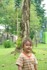 A cheerful young girl stands in a lush park, smiling at the camera. Surrounded by trees and green grass, the scene conveys a warm, carefree moment outdoors.