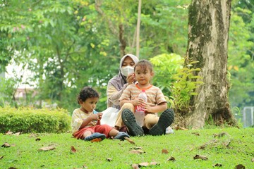 Fototapeta premium A mother and two children relax on a sunny park lawn, sharing a playful moment outdoors. The scene captures family bonding, nature, and everyday happiness in a green urban park.