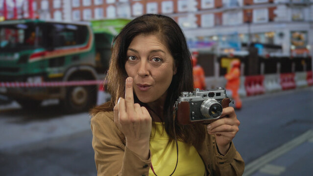 Woman holding vintage camera making middlefinger gesture in front of building; bold defiance attitude.