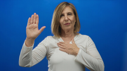 Fototapeta premium Middle-aged caucasian woman raising palm and placing hand on chest in studio against blue wall; sincerity.