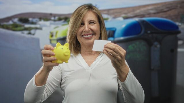 Middle age woman holds yellow piggy bank and blank card by recycling container on street; financial security confidence.