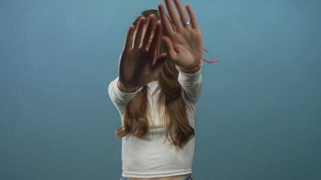 Young hispanic woman wearing white long sleeve top holding palms out to block camera in blue studio, visible rings and red bracelet; shyness. - Powered by Adobe