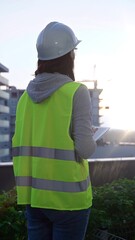 Woman construction engineer with a white hard hat and safety vest is using digital tablet while inspecting a construction site in the early morning at sunrise, back view