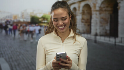 Woman smiling holding smartphone in front of roman coliseum in italy outdoors surrounded by...