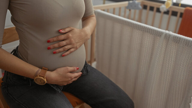 Pregnant woman in a bedroom gently holds her belly near a baby crib, showcasing maternal anticipation and warmth in a serene setting. - Powered by Adobe