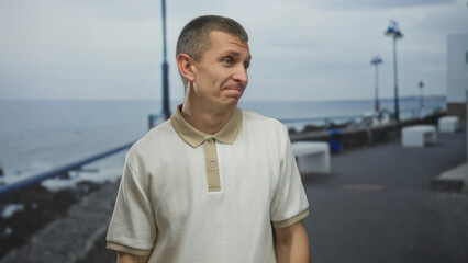 Man stands on a seaside promenade pointing sideways wearing casual polo with overcast sky in the background, giving a relaxed gesture near the beach.