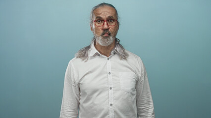 Fototapeta premium Man with long gray hair and beard wearing red glasses and white shirt purses lips in light blue studio; surprise and quiet introspection.