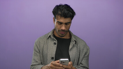 Fototapeta premium Man scrolling smartphone with both hands and downward gaze in studio with purple background; digital concentration.