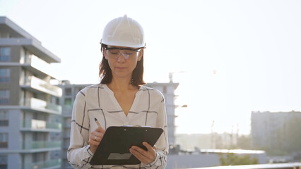 Woman construction engineer wearing white checked blouse and hard hat is making notes on a clipboard while inspecting a building site at sunset, front view. Architecture and engineering concepts