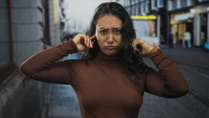 Woman in brown turtleneck plugging her ears with fingers and frowning on a busy street with tram...