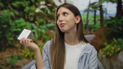 Woman holds creditcard with outstretched hand at forest clearing under vibrant green foliage;...