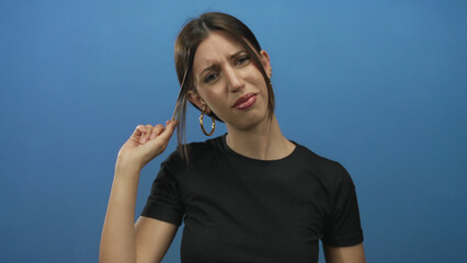 Woman holds a hair strand between fingertips in a blue studio setting with a puzzled frown;...