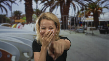 Blonde woman covers bare cheek with hand while pointing finger at camera on busy street lined with...