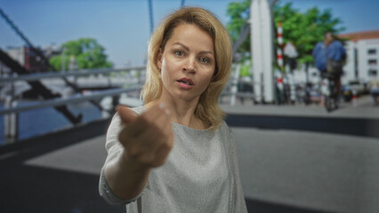 Woman in gray blouse standing on urban street beside canal railing points finger; assertion confidence.