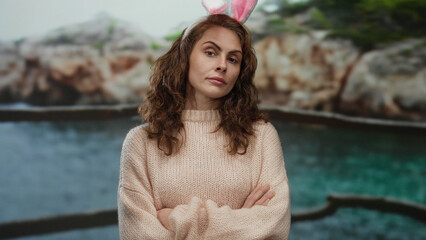 Woman with bunny ears at the beach crossing arms in front of the sea under an overcast sky next to...