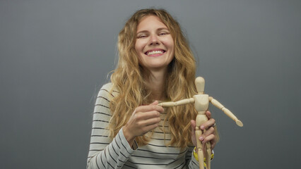 Woman holds mannequin while smiling in studio with blonde young hair framing a wooden posing figure and adjusting its casual posture.