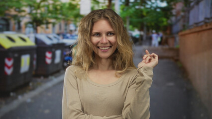 Fototapeta premium Woman lifts hand and presents a gentle smile on a street while blonde young hair flows against a city backdrop highlighting her outdoor expression.