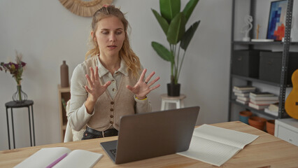 Woman with a laptop typing from home tilts her head behind blonde young locks while sitting at a desk in a modern interior remote workspace.