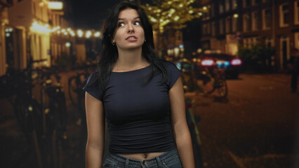 Dark-haired woman wearing navy crop top looks up with slight head tilt on street under warm lights; quiet contemplation.