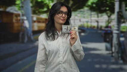 Woman wearing glasses holding hundred dollar bill on street paved sidewalk under green trees; wealth confidence.