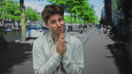 Man with hands together in prayer on an amsterdam street, eyes closed and pleading expression;...