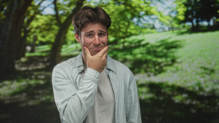 Young hispanic man with hand on cheek for toothache in green park under trees and sunlight; pain...