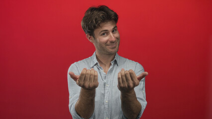 Man young hispanic offering both cupped hands palms forward with slight smile, wearing striped button shirt in red studio; invitation welcome playful.