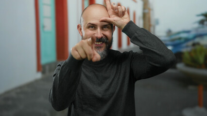 Obraz premium Bearded man gesturing loser sign with hand on forehead in urban street setting, showcasing humor and confidence.
