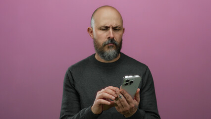 Bald man with beard using smartphone against isolated pink background with focused expression and...