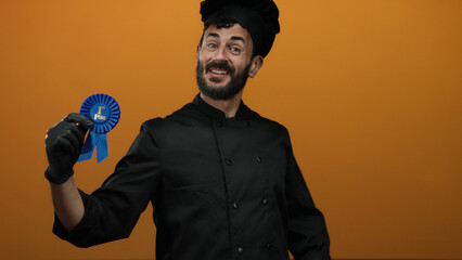 Hispanic man wearing chef uniform holding first place ribbon, smiling against vibrant orange background, showcasing his culinary achievement.