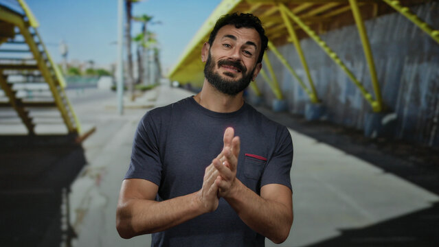 Hispanic man with beard clapping hands outdoors on a sunny street under yellow structure showing contentment and casual attitude.
