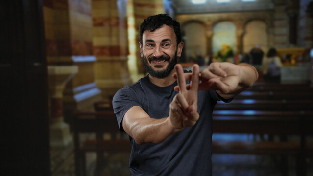 Hispanic man gesturing hashtag with fingers inside a catholic church, surrounded by wooden benches and vibrant architecture, capturing a moment of modern expression in a traditional setting.