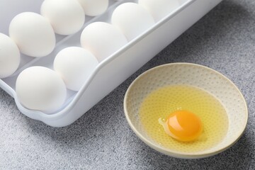 Many whole raw chicken eggs and broken one in bowl on grey table, closeup
