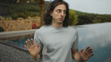 Man with long hair points finger at pool beside hotel building outdoors while looking sideways; awkward surprise.