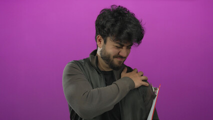 Young man with beard holding shoulder against pink background, implying discomfort or pain, wearing...