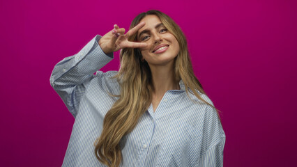 Fototapeta premium Woman making peace sign with hand in a bright purple photo studio showcasing her long blonde hair; joy playfulness confidence positivity.