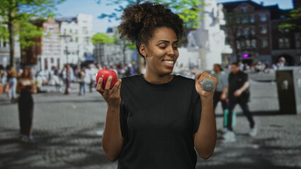Woman holding apple and dumbbell while smiling on busy street plaza with buildings and trees...