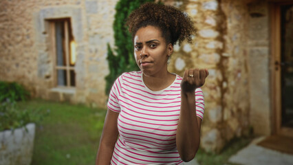 Fototapeta premium Woman in pink striped t shirt holds clenched fist near shoulder and lifts other hand palm up at a stone building entrance outdoors; frustration.