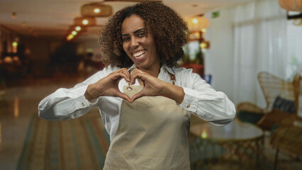 Young woman baker wearing apron in hotel lobby forms heart with hands while smiling broadly and looking at camera; joy hospitality welcome.