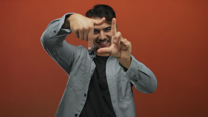 Fototapeta premium Young hispanic man making frame gestures with hands on an isolated red background, expressing creativity and focus in a filmmaking or photography context.