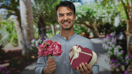 Young man holds flowers and heart-shaped gift box in a vibrant park setting under sunny skies,...