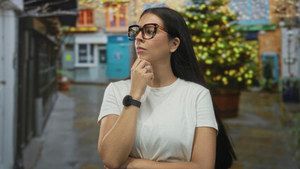 Woman with glasses and hand on chin in a street alley by a planter and building, wearing white t shirt and watch; thoughtful reflection.