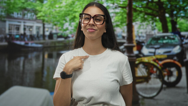 Woman hand on chest wearing oversized glasses and white t shirt with ring and smartwatch on street by canal in amsterdam; calm contentment.