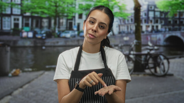 Woman in apron pinches fingers to palm on street by canal and building, showing hands and gesture; confidence teaching.