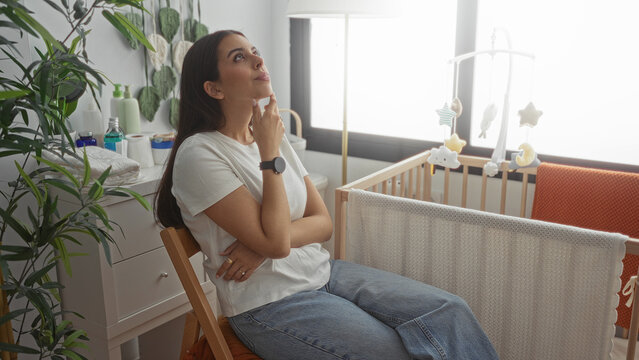 Woman sitting on chair with hand to face and arms folded beside wooden baby crib and hanging mobile in building; exhaustion new parent.